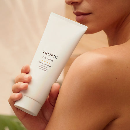 The image features a tube of Tropic Body Love Firming Buttercream, positioned against a textured, sunlit background. Key benefits of the product are highlighted:
Zingy citrus scent, offering a refreshing and uplifting fragrance.
Toning, designed to help firm and tighten the skin.
Hydrating, enriched with ingredients like shea butter, pink peppercorn, and vitamin E to deeply moisturize.